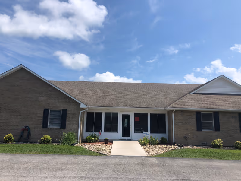 Front exterior view of a single-story brick building with a gable roof under a partly cloudy blue sky. There is a concrete walkway leading to a glass door entrance flanked by windows. Small shrubs and plants are arranged along the front of the building.