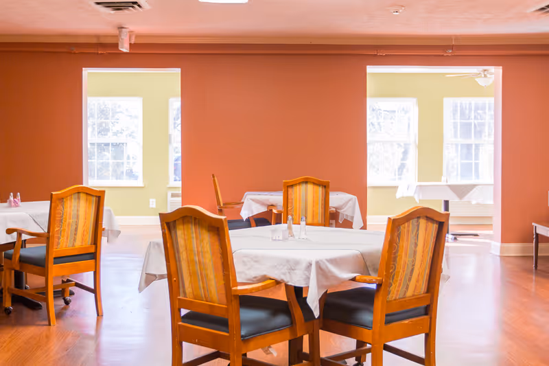 A bright dining room with round tables covered in white tablecloths and wooden chairs against warm-colored walls and large windows.