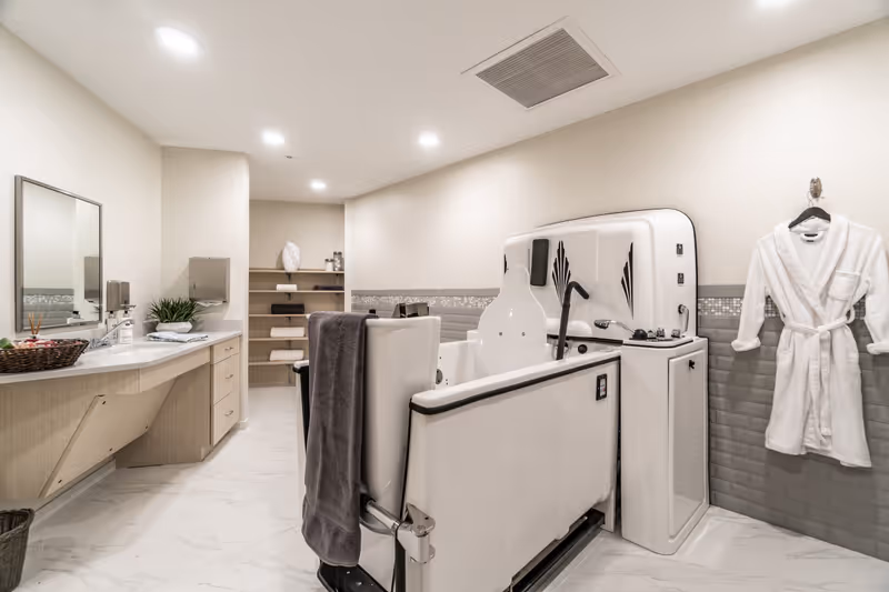 A spacious, well-lit bathroom with a large accessible bathtub equipped with safety handles and controls. A white bathrobe hangs on the wall to the right. On the left side, there is a long countertop with a sink, a mirror, a basket with toiletries, and a paper towel dispenser. Shelves with neatly folded towels and other items are visible in the background. The floor is tiled in white, and the walls are painted in light neutral tones with a decorative tile border.
