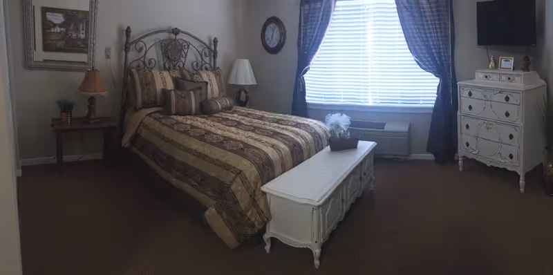 A cozy bedroom with a decorative metal headboard bed covered in a striped and floral patterned comforter with multiple pillows. There is a white wooden chest at the foot of the bed, a small wooden side table with a lamp and plant on the left, and a white dresser with a TV mounted above it on the right. A window with blinds and dark curtains lets in natural light.