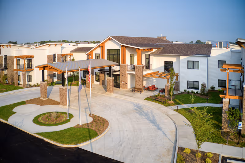 Exterior view of Preston Greens Senior Living facility showing a modern two-story building with white siding, brick accents, and wooden beams. The entrance features a covered drop-off area with three flagpoles and landscaped greenery around the driveway.