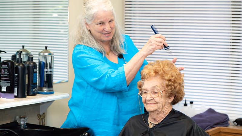 An elderly woman with curly hair and glasses is sitting in a salon chair wearing a black cape while another woman with long white hair and a blue shirt is styling her hair with a comb. Various hair care products are visible on a counter beside them, and a window with blinds is in the background.