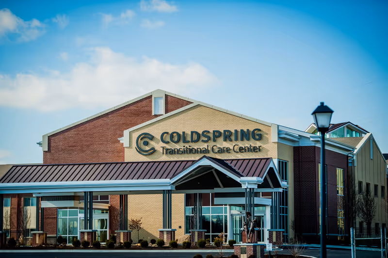 Exterior view of Coldspring Transitional Care Center building with a covered entrance, brick and beige walls, large windows, and a street lamp in front under a partly cloudy blue sky.