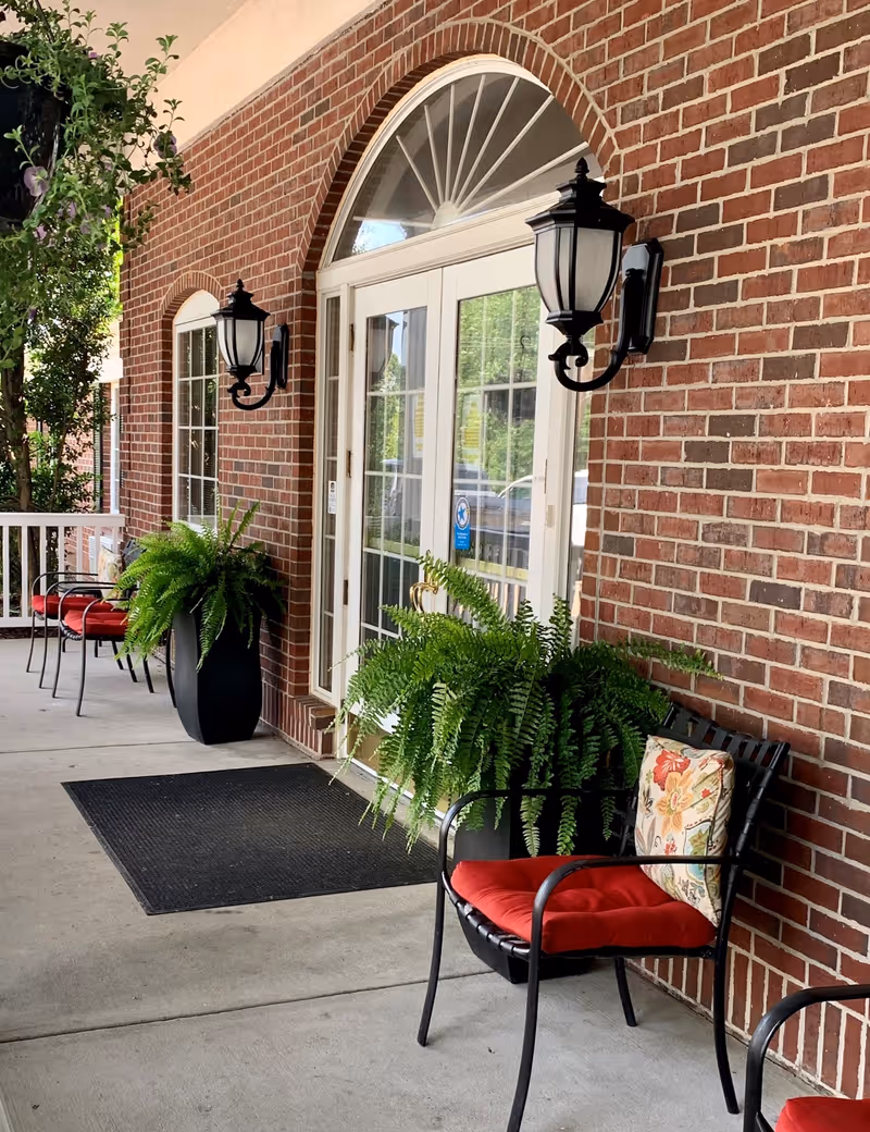 Outdoor entrance area of The Stilley House Senior Living featuring a brick wall with white-framed glass double doors and arched window above. Two black wall-mounted lanterns flank the doors. Black metal chairs with red cushions and floral pillows are arranged along the concrete porch, accompanied by large potted green ferns. A black doormat is placed in front of the doors.