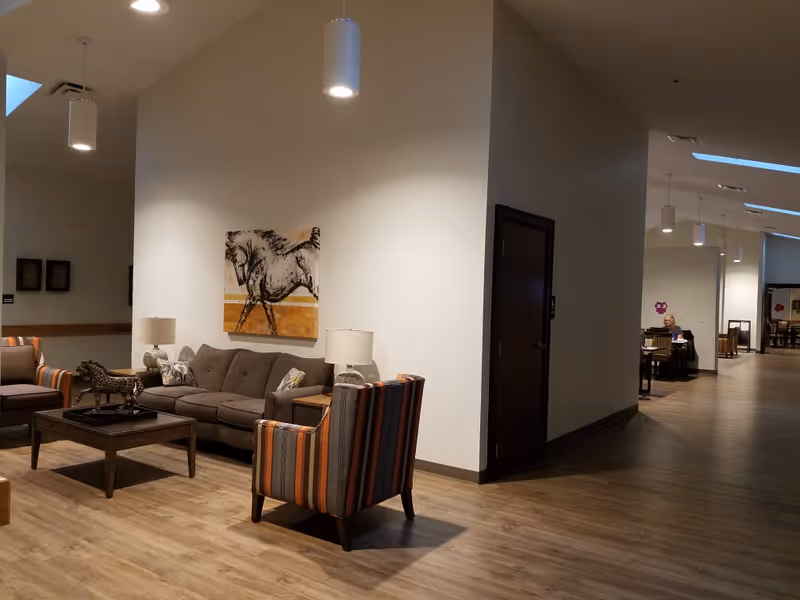 Communal seating area with a sofa, striped armchairs, coffee table, lamps, and wall art in a senior living facility hallway.