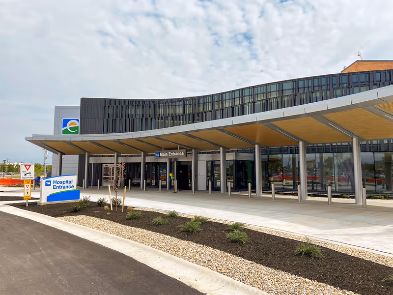 Exterior view of St. Elizabeth Hospital Florence showing the main entrance with a covered walkway. There is a sign indicating the hospital entrance and a pedestrian yield sign near the driveway. The building has large glass windows and a modern design under a partly cloudy sky.