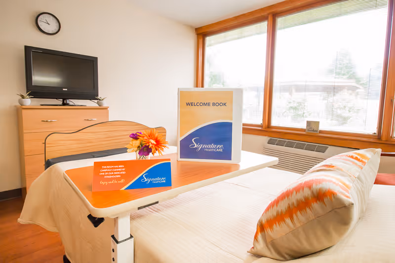 Sunlit resident bedroom with a made bed, an overbed table holding a Welcome Book and flowers, a TV on a dresser, and large windows.