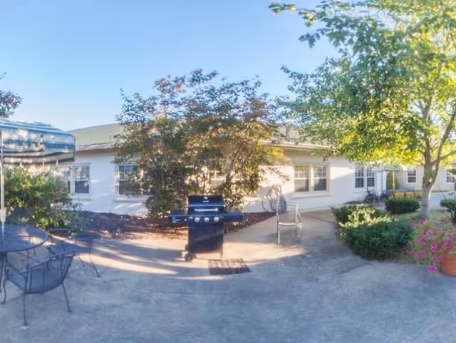 Outdoor patio area at Crestview Healthcare and Rehabilitation featuring a black grill, metal chairs, a table, and surrounding greenery including trees and bushes under a clear blue sky.