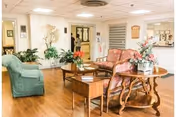 A nursing home common area with wooden flooring, featuring a green sofa, two floral upholstered armchairs, a wooden coffee table with a flower arrangement, and additional plants and decorations in the background.
