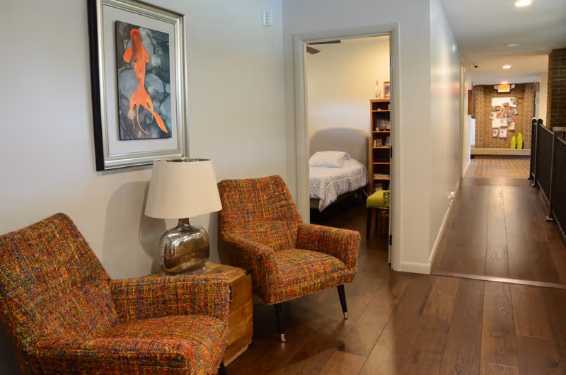 A hallway in Southern Grace Assisted Living with two colorful armchairs and a lamp on a wooden side table. A framed picture of an orange fish hangs on the wall above the chairs. The hallway leads to a bedroom with a bed covered in a blue and white patterned bedspread and a wooden bookshelf. The floor is dark wood, and the hallway ends with a brick wall and an exit sign.