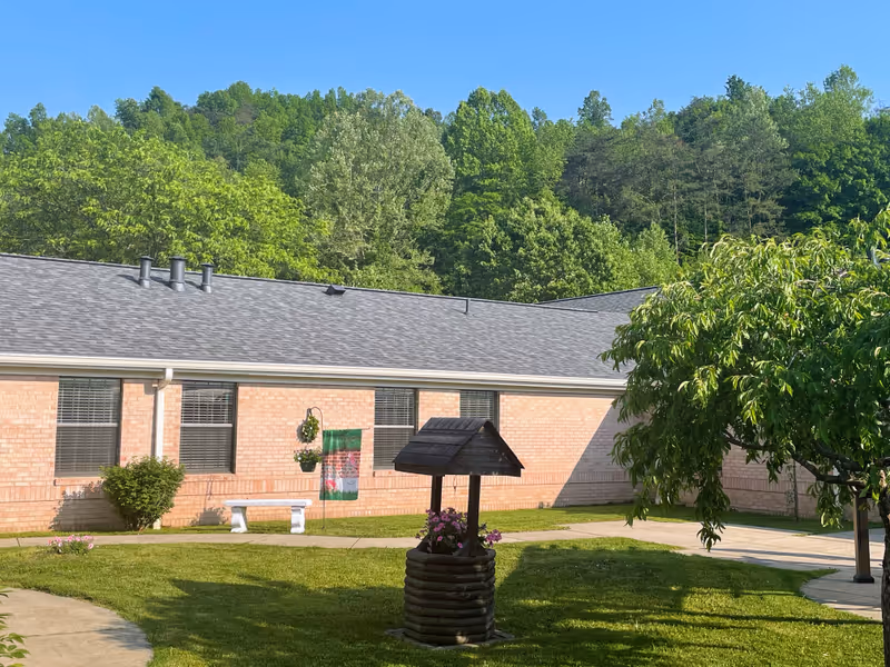 Outdoor courtyard area of Elliott Nursing and Rehabilitation with a brick building in the background, green grass, a small wooden wishing well planter with flowers, a white bench, a hanging flower basket, and a tree. Trees and a clear blue sky are visible beyond the building.