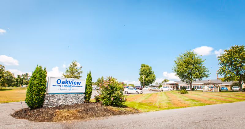 Exterior view of Oakview Nursing & Rehabilitation Center with its entrance sign, parking lot, lawn, and building under a blue sky.
