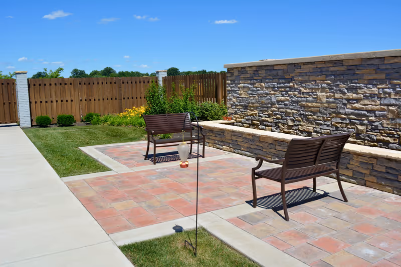 Outdoor brick patio with two benches, lawn, and stone wall under a blue sky.