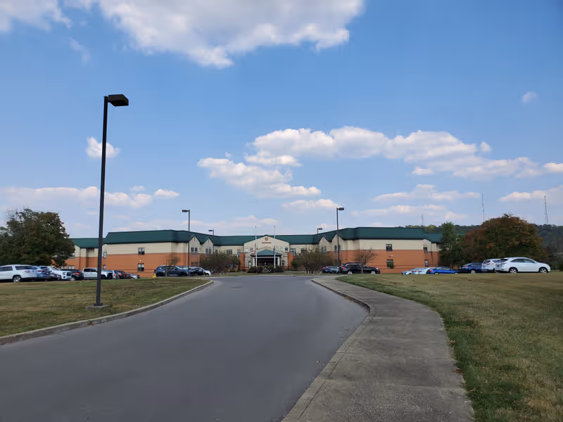 Senior living facility building with a curved driveway, parking lot, and green lawn under blue sky.