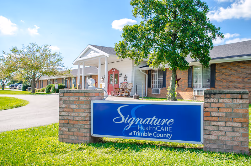 Front exterior of a single-story brick senior living facility with a blue 'Signature HealthCARE of Trimble County' sign on the lawn.