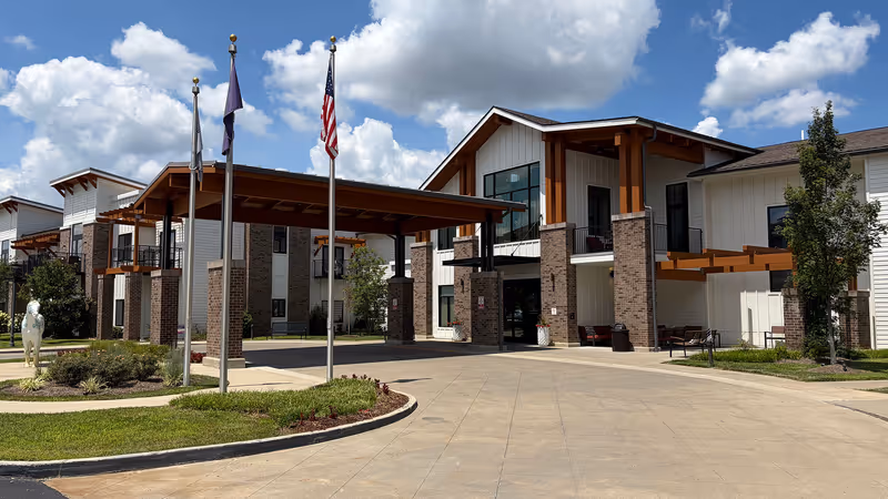 Modern senior living facility exterior with brick pillars, a porte-cochère, and flagpoles.