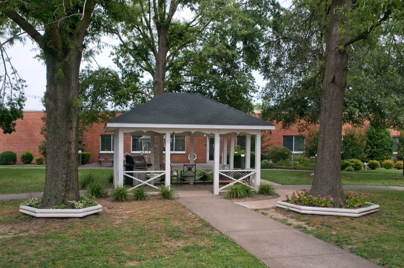 A small white gazebo with a dark shingled roof is situated in a grassy courtyard with two large trees on either side. There are benches and a grill inside the gazebo. In the background, a red brick building with windows and a door is visible, surrounded by neatly trimmed bushes and landscaping.