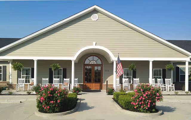 Front exterior view of a single-story senior living facility building with beige siding, a central entrance with a wooden door and arched window above it, white columns supporting a covered porch, several white rocking chairs on the porch, hanging plants, an American flag near the entrance, and landscaped bushes with pink flowers in front.