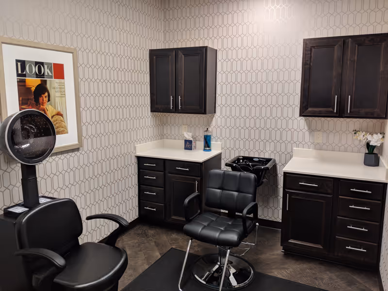 Hair salon area with dark wood cabinets, black styling chairs, a hooded dryer, and geometric wallpaper.