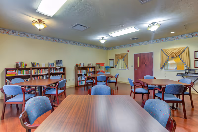 A senior living facility common area with tables, blue chairs, and bookshelves.