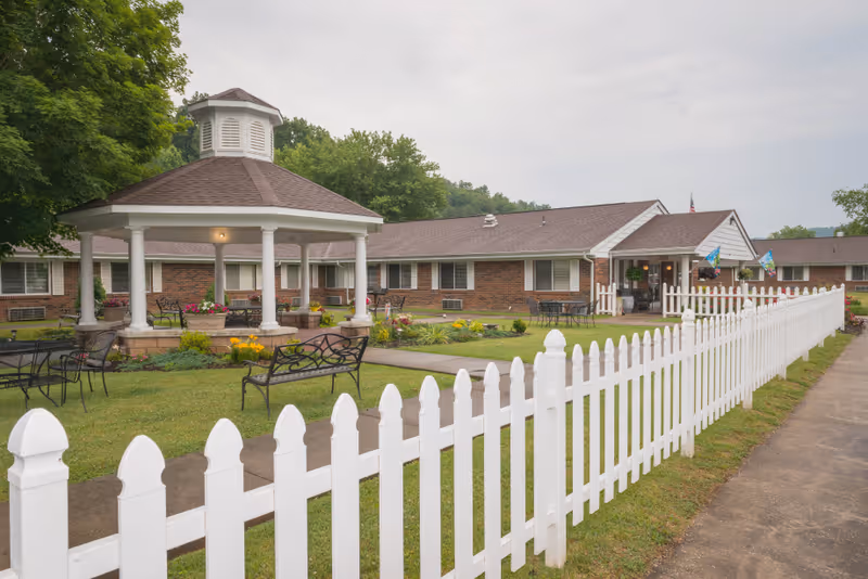 Front lawn of a senior living facility featuring a white picket fence, a gazebo with benches, flowerbeds, and a low brick building with an entrance.