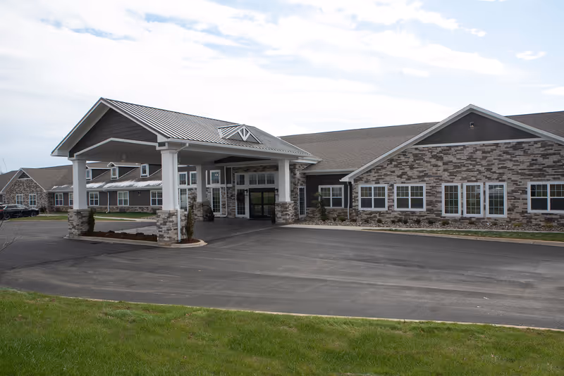 Exterior view of a senior living facility building with a covered entrance supported by white columns and stone accents. The building has multiple windows and a combination of stone and siding on the facade. There is a paved driveway and a grassy area in the foreground under a partly cloudy sky.