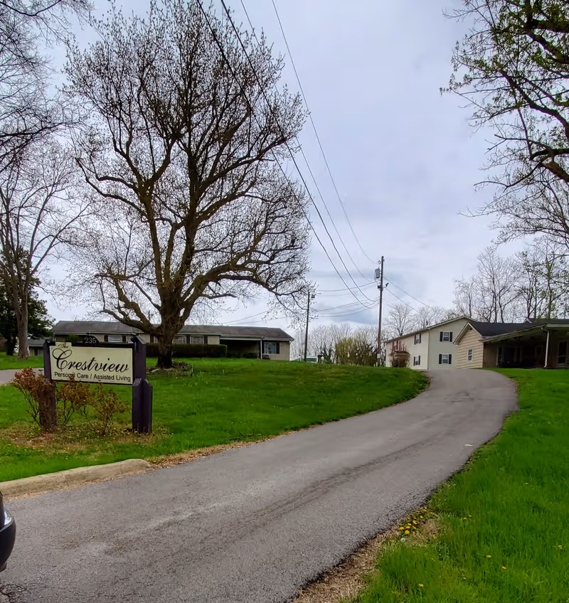 A rural road curves toward Crestview Personal Care facility with a sign and trees.