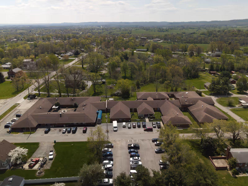 An aerial view of a large brown-roofed care facility with parking and trees.