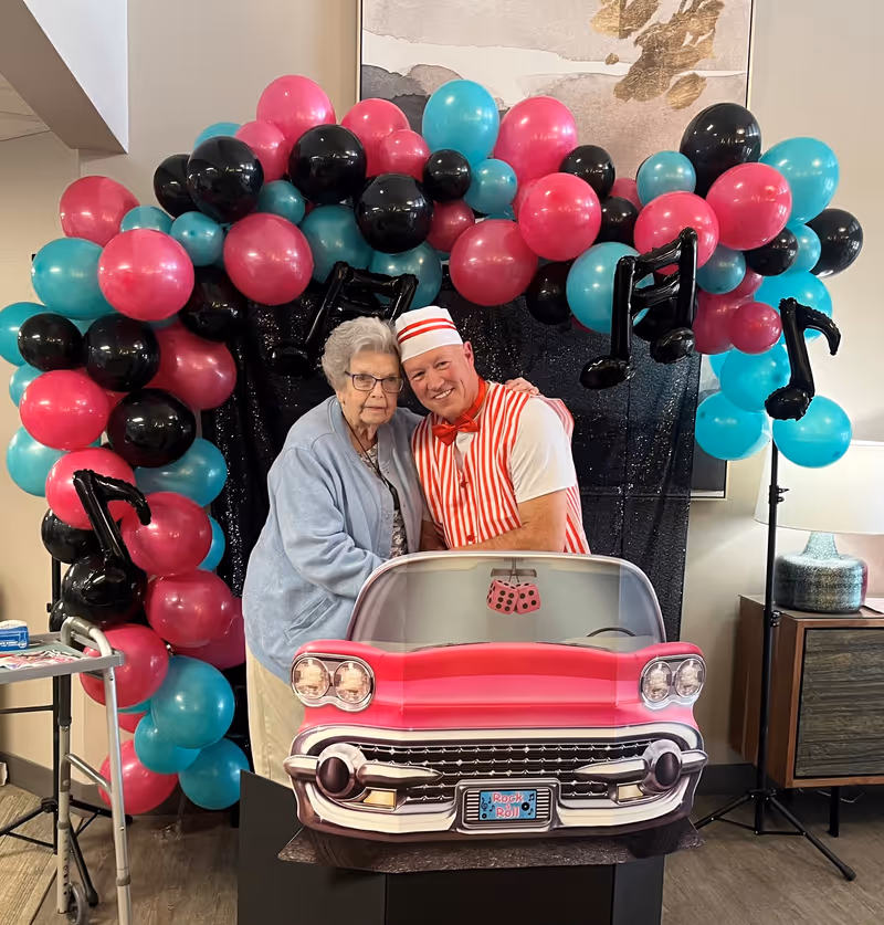 An elderly woman and a man dressed in a red and white striped outfit with a matching hat pose together behind a pink vintage car photo prop. They are standing in front of a colorful balloon arch made of pink, black, and teal balloons, with black musical note balloons integrated into the arch. The setting appears to be indoors with a walker and a side table with a lamp visible nearby.