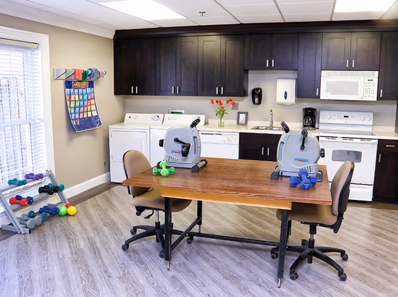 A room with a kitchen area featuring dark wood cabinets, a white stove, microwave, dishwasher, and washer and dryer. In front of the kitchen is a wooden table with two small exercise bikes and dumbbells on it. Two office chairs are positioned at the table. To the left, there is a rack with various dumbbells and colorful balls, and a calendar hanging on the wall near a window with blinds.