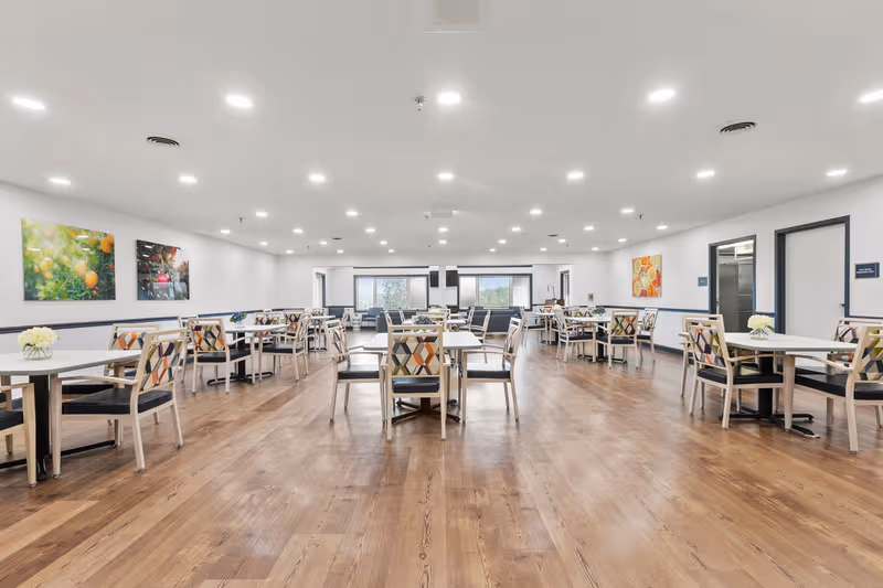 Spacious dining room with multiple tables and chairs, bright overhead lighting, and wall art.