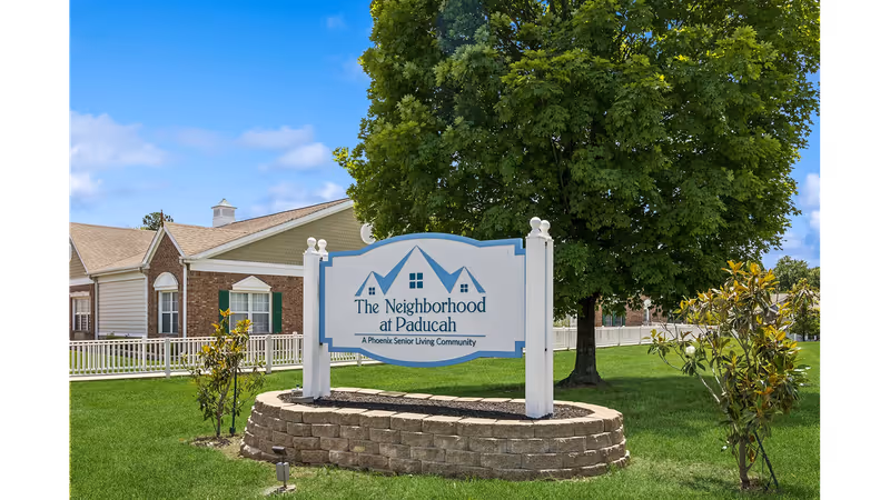 A white and blue entrance sign reading "The Neighborhood at Paducah" on a raised stone bed with a large tree and senior living buildings in the background.