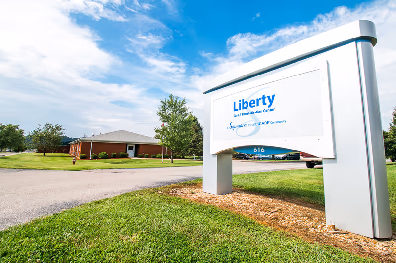 Outdoor view of Liberty Care and Rehabilitation Center sign with a building and parking lot in the background under a partly cloudy blue sky.