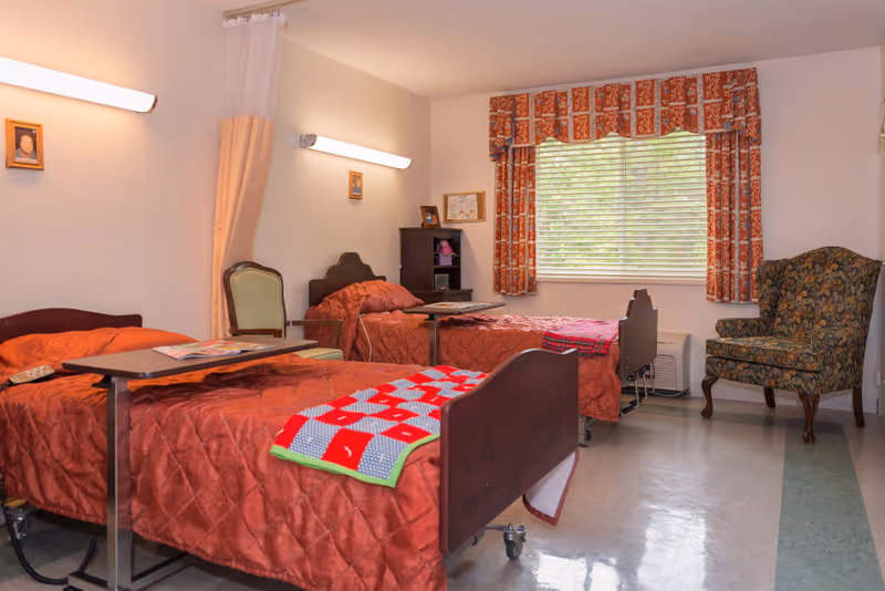 A room with two hospital-style beds covered in red quilts, each with an overbed table. There is a green upholstered chair between the beds and a floral armchair near a window with patterned curtains. The room has beige walls and fluorescent wall lights above each bed.
