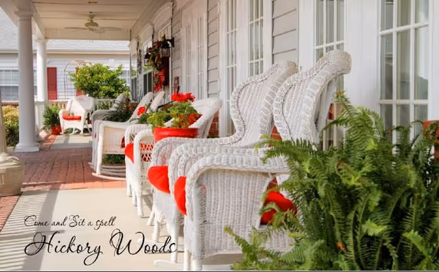 A porch with white wicker chairs lined up along the side of a building with large windows. The chairs have red cushions and there are potted plants and flowers placed on and around the chairs. The porch has a brick floor and white columns supporting the roof.