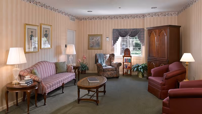 A cozy living room with a striped sofa, two red armchairs, a floral armchair, wooden coffee table, side tables with lamps, framed artwork on the walls, a window with a valance, and a wooden cabinet. There are also some plants and books on a small shelf near the window.