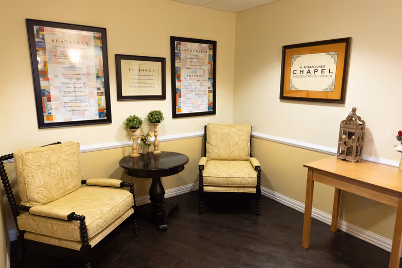 A small, cozy corner room with two cushioned armchairs upholstered in yellow patterned fabric, a round dark wooden table between them holding two small potted plants and a small flower arrangement. The walls are painted beige with white trim and decorated with framed plaques and signs, including one that reads 'M. Susan Jones Chapel and Education Center.' A wooden table with a decorative lantern and a vase with flowers is positioned against the right wall. The floor is dark wood.