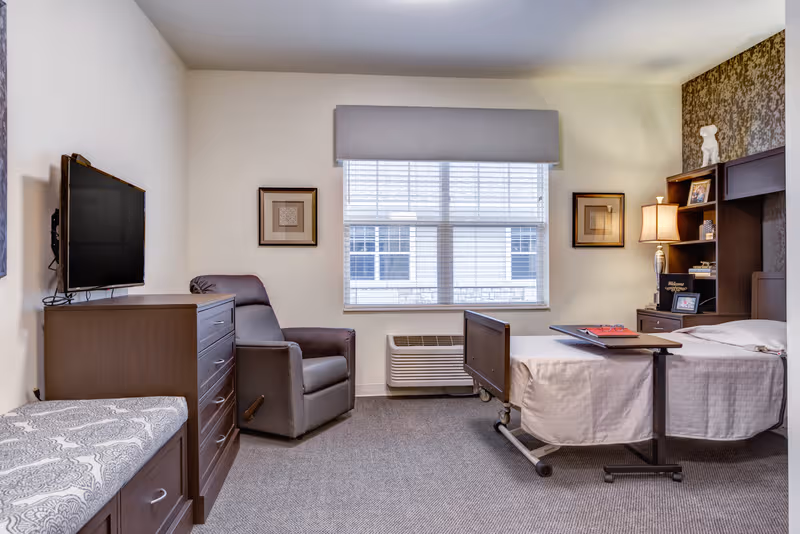 A senior living bedroom with bed, recliner, dresser, TV, and window.
