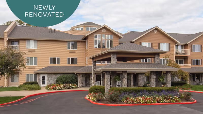 Exterior view of a newly renovated multi-story senior living facility with beige siding and stone accents. The building features multiple windows, a covered entrance with stone pillars, and landscaped flower beds surrounding the driveway.