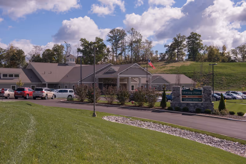 Exterior view of The Lantern at Morning Pointe Alzheimer's Center of Excellence in Russell, showing a single-story building with a pitched roof, surrounded by a parking lot with several cars, green grassy areas, trees in the background, and a partly cloudy sky.
