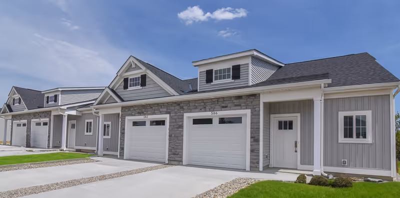 Exterior view of a modern residential building with multiple attached units, each featuring a white garage door and a front entrance. The building has gray siding with stone accents and black shutters on the upper windows. The driveway and small patches of green lawn are visible under a partly cloudy blue sky.