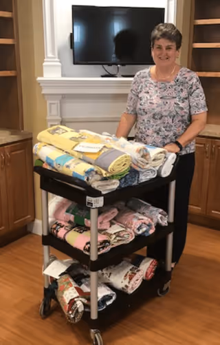 A woman standing behind a black three-tiered cart filled with neatly folded colorful quilts in a room with wooden cabinets and a mounted flat-screen TV above a white fireplace mantel.