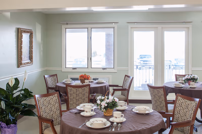 Bright dining room with round tables set with china and floral centerpieces, upholstered chairs, and large windows and doors.
