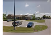 Single-story brick care center building with a front parking lot, several parked cars, light posts, and grassy islands under a partly cloudy sky.