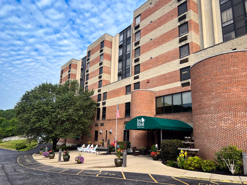 Exterior view of a multi-story brick building with a green awning entrance labeled 'Ivy Knoll'. There are potted plants and flowers near the entrance, a flagpole with an American flag, outdoor seating with white chairs, and a large tree to the left. The sky is partly cloudy.