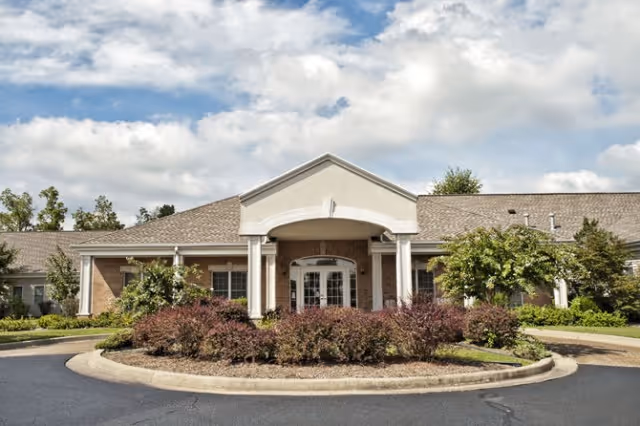 Front entrance of a single-story senior living building with a covered portico, columns, circular driveway, and landscaped shrubs.