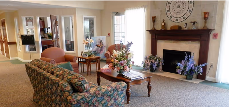 Carpeted senior living room lounge with a floral sofa and armchairs around a coffee table topped with flower arrangements in front of a fireplace.