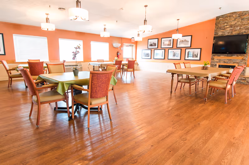 A bright dining room with wooden floors, several tables covered with tablecloths, and chairs arranged around them. The walls are painted orange and decorated with framed pictures. There are large windows letting in natural light and a stone fireplace with a mounted flat-screen TV.