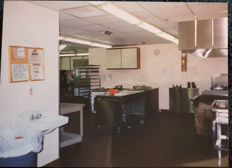 Industrial kitchen inside a facility with stainless counters, shelves, and a large vent hood.
