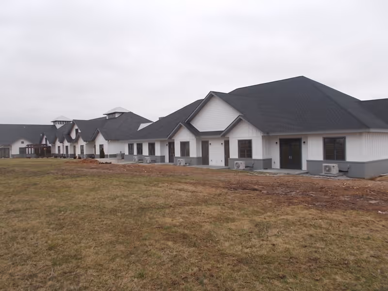 Exterior view of a large, single-story assisted living facility building with white siding and dark gray roofing, situated on a grassy area under an overcast sky.